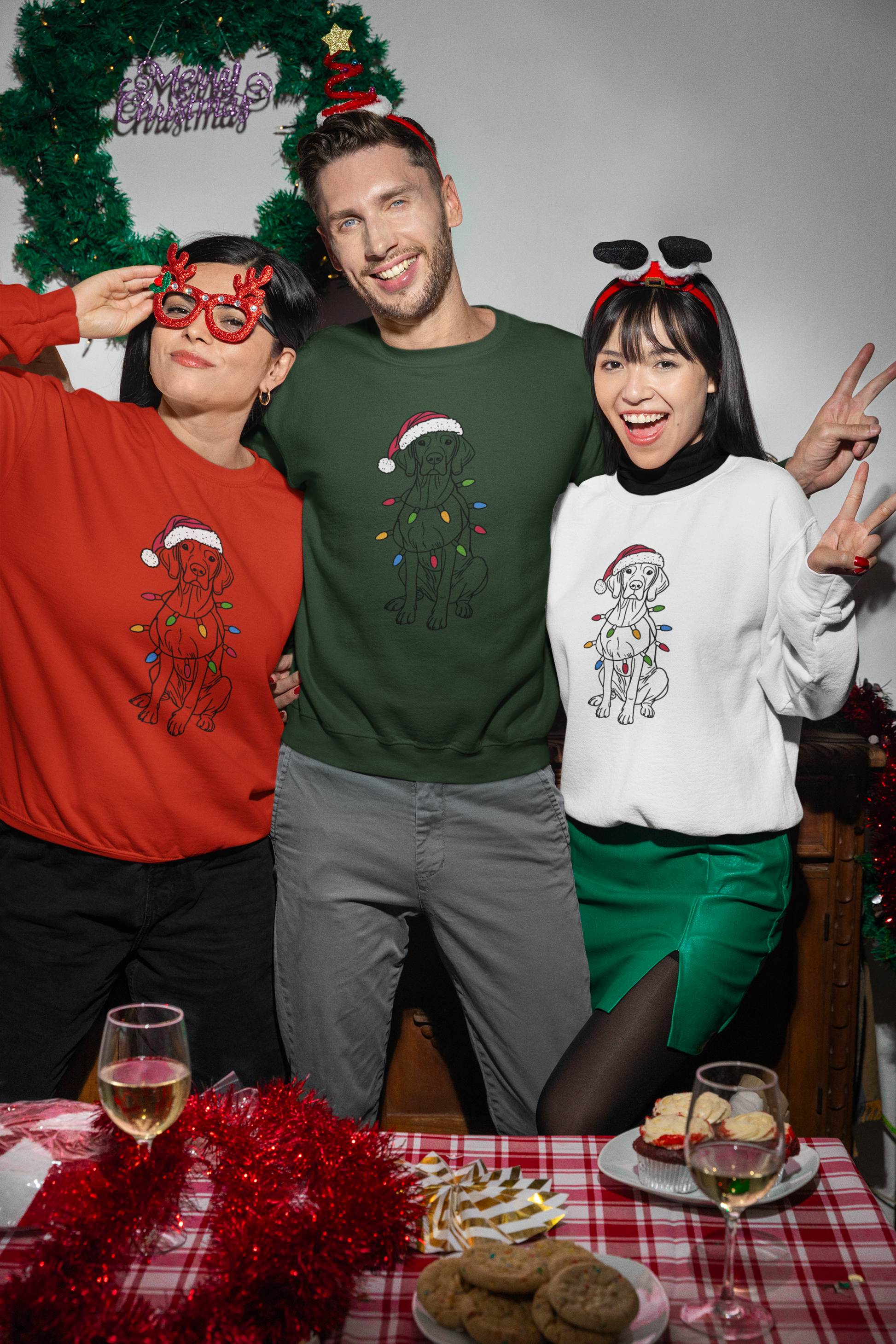 two woman and a man wearing red green and white christmas sweaters with a vizsla on it wearing a christmas hat and christmas lights