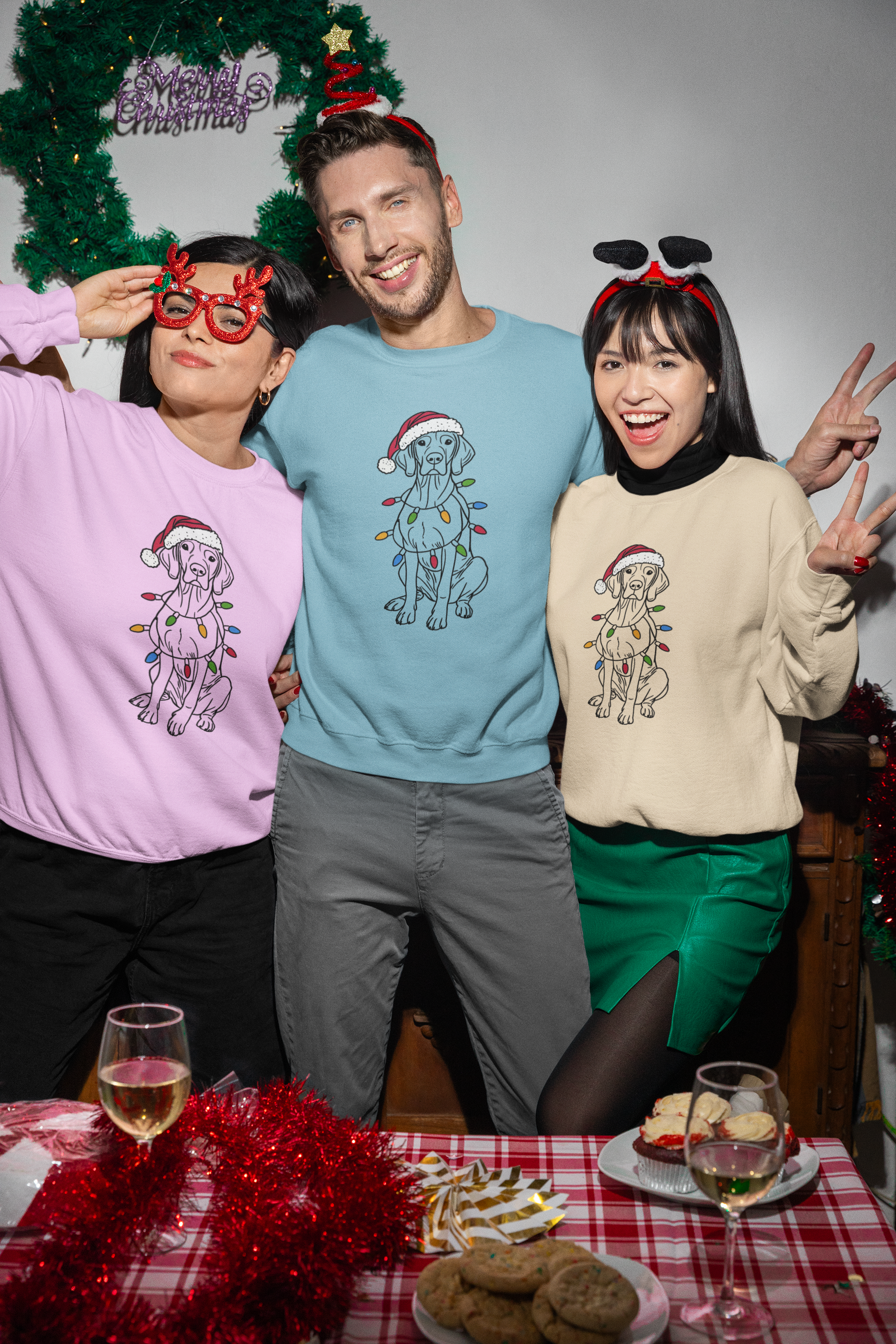 two woman and a man wearing pink light blue and beige christmas sweaters with a vizsla on it wearing a christmas hat and christmas lights